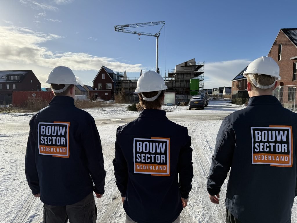 Drie vakmensen van Bouwsector Nederland met witte helm op een besneeuwde straat met woningen in aanbouw